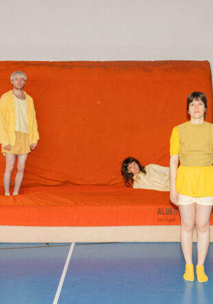 Trois personnes habillées en jaune qui sont dans une salle de gymnastique au sol bleu et qui posent autour, devant et sur des grands tappis de gymnastique orange très épais. coucou studio