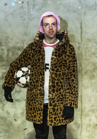 Félicien est pris en photo couleur avec le maquillage effacé d'un clown triste. Il porte un manteau de fourrure léopard, un bob rose et il tient un ballon de football sous le bras droit. Giona Mottura & Paola Ghiano