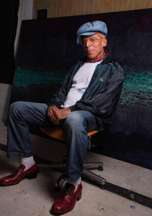 Satch Hoyt qui pose assis devant une oeuvre peinte. 
