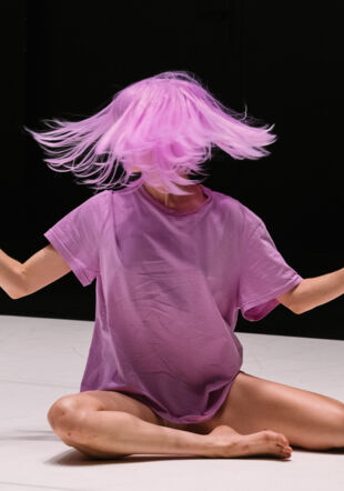 L'artiste est assise en tailleur avec les bras à mis hauteur écartés. Elle porte un t-shirt large de couleur Lilas et une perruque de la même couleur qu'elle secoue de droite à gauche. M. Magnin