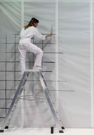 L'artiste habillée tout en blanc se trouve en haut d'une échelle. On la voit peindre sur un mur recouvert de protections blanches. Elle peint un quadrillage noir. Jean-Louis Fernandez