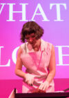 Jeanne Spaeter habillée d'un tablier blanc et d'une chemise blanche  découpe une coeur sur une planche en bois en portant des gants en latex blancs. Derrière elle une image rose/ violette est projetée avec la phrase "What is love?" Elisabeth Blaettler
