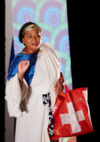 Il s'agit d'une photo de l'artiste en couleurs qui porte un drapé blanc au-dessus d'un tissu bleu brillant. Elle porte un sac de commission au couleur du drapeau suisse. Elle semble être entrain de s'exprimer et utilise son bras et sa main dans un geste e Ronja Burkard