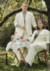 Les deux artiste posent dans un jardin verdoyant, é'une assise sur une chaise. l'autre debout. Il y a une petite table ronde avec une nappe blanche avec des petites choses à manger. Les deux personnes sont habillées en costumes blancs. 